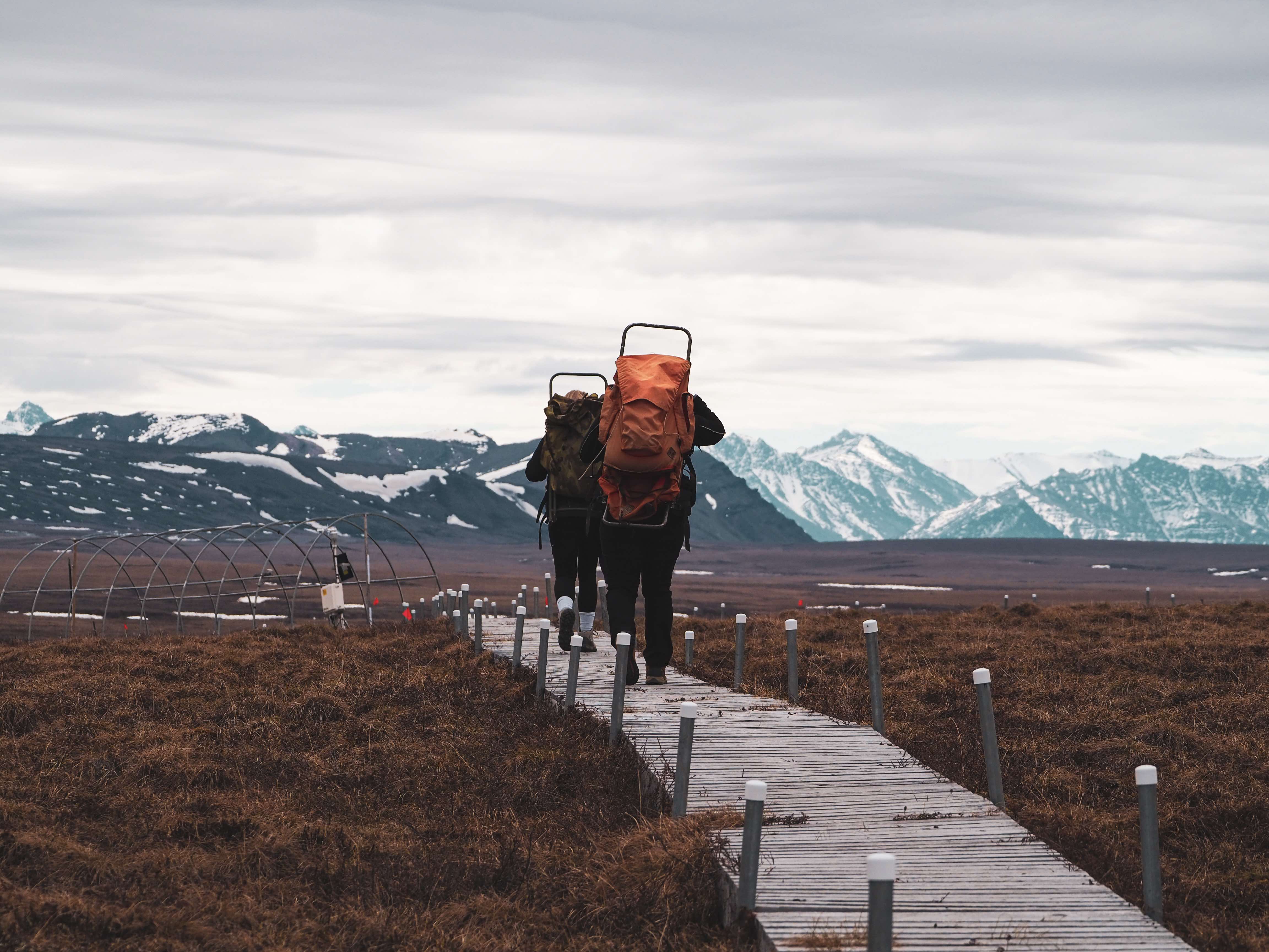 Photo of two people walking away from the camera on a small boardwalk with large backpacks while mountains tower in the background